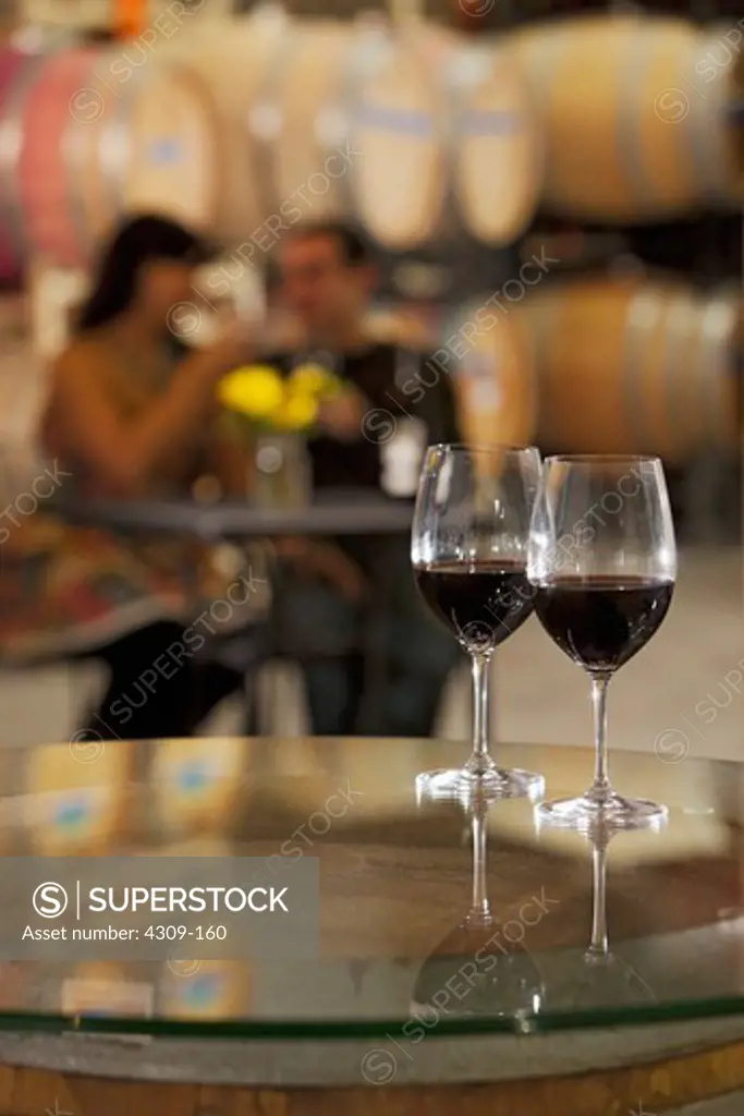Two glasses of red wine on a table, with a couple tasting wine in the background.