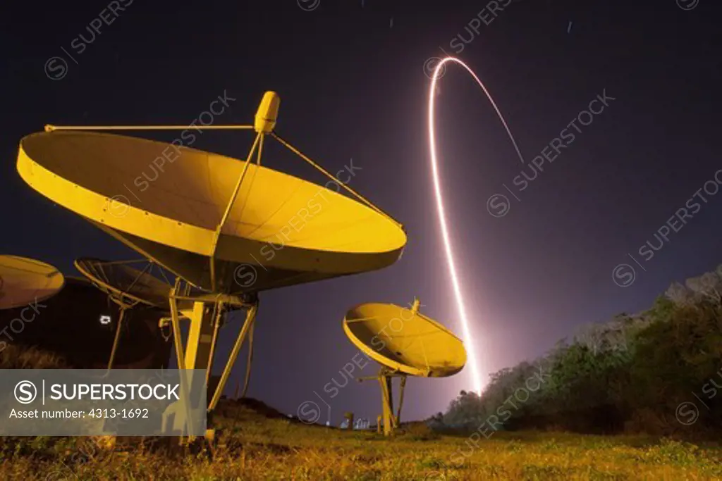 USA, Florida, Cape Canaveral, Kennedy Space Center, view of satellites and space rocket launch at night. A time-lapse streak photograph captures the path of an Atlas V (Atlas 5) rocket as it launches NASA's Tracking & Data Relay System satellite, TDRS-K, into space. In the foreground are satellite dish antennas.