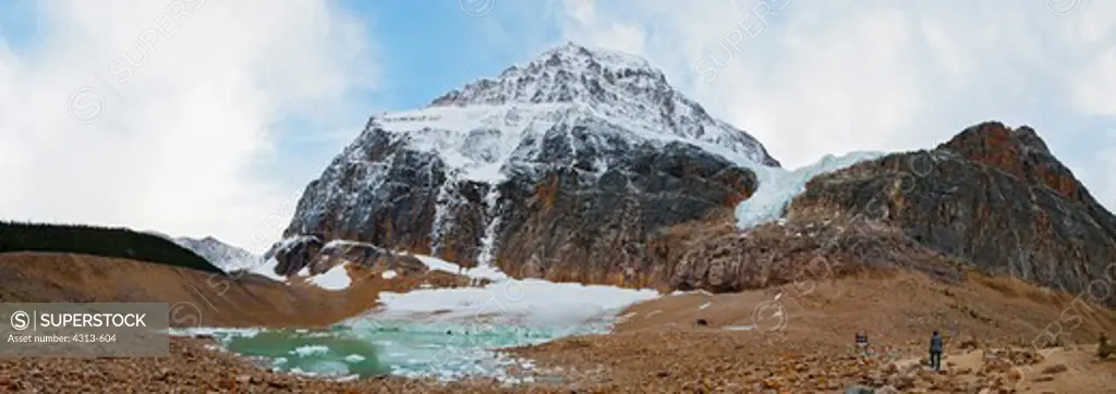 A panorama shows a hiker standing at the base of Mount Edith Cavell, where fast-melting Angel and Cavell glaciers shed icebergs into Cavell Pond. The entire valley here was covered by the glacier as recently as just a few decades ago.