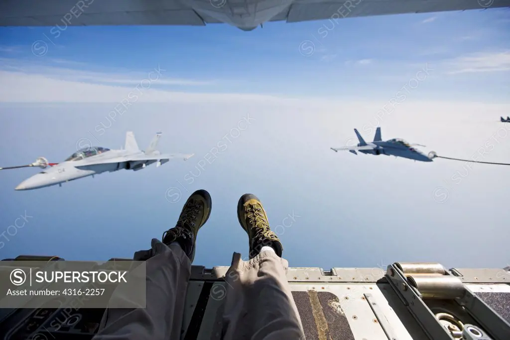 A view of a photographer's legs and feet dangling above the South China Sea from a United States Marine Corps KC-130J as it refuels two F/A-18D Hornets.