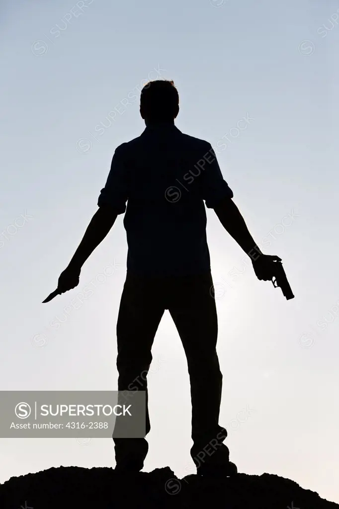 An adult male holds a handgun in one hand and a fighting knife in the other hand.