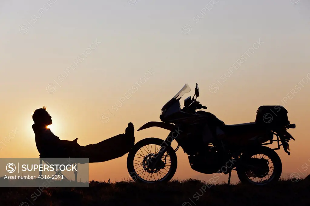 A man at a camp in the mountains relaxes with his feet up against his off road motorcycle.