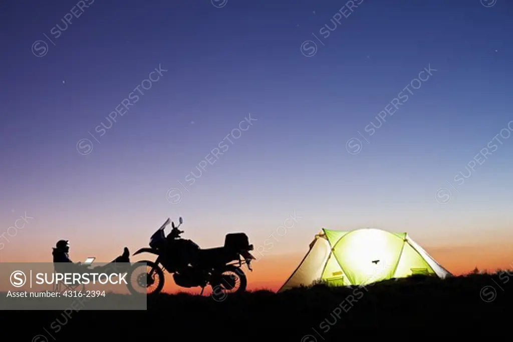 A man working on a laptop computer with his feet up against his motorcycle at his camp at dusk.