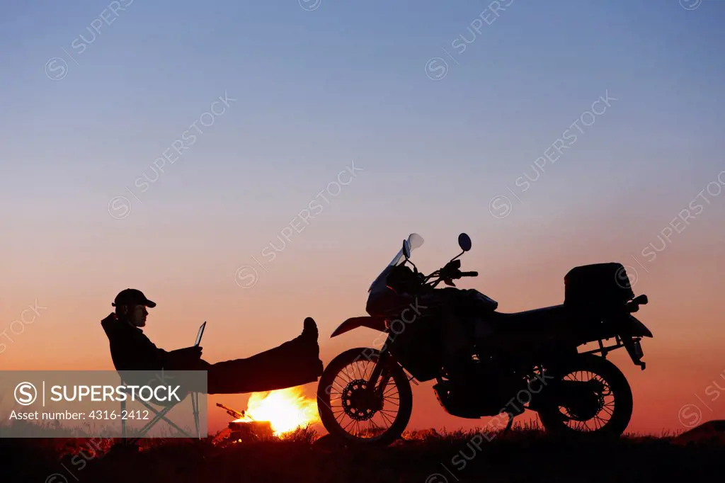 With a campfire burning, a man works on his small laptop computer at dusk as he rests his feet on the front wheel of his off road motorcycle.