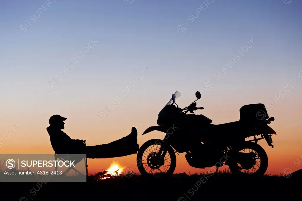 As a campfire burns, a man relaxes at his camp with his feet against the front wheel of his off road motorcycle.