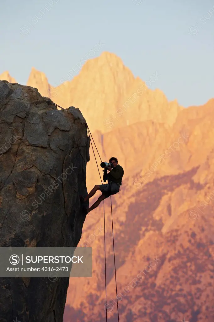 A photographer, suspended by a rope on the side of a cliff, using a long telephoto lens to photograph, is partially silhouetted by sunlight on the face of Mount Whitney and surrounding peaks. The photographer is in the Alabama Hills of California, near the town of Lone Pine.