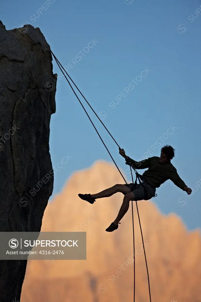 A climber swings out while rappelling off of a cliff , silhouetted against Mount Whitney in California's eastern high Sierra Nevada mountains. The climber is in the Alabama Hills, near the town of Lone Pine.