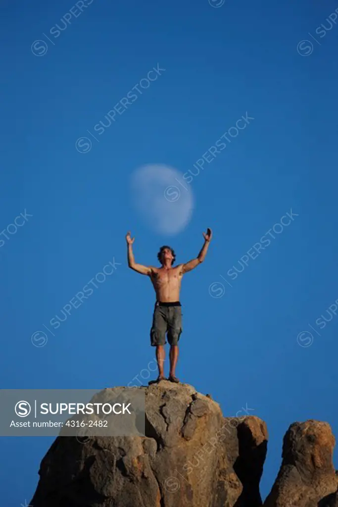 A climber, standing atop a boulder with his arms outstretched, appears to be grasping a gibbous moon rising above the Alabama Hills of California, near the town of Lone Pine, below Mount Whitney and the high Sierra Nevada Mountains
