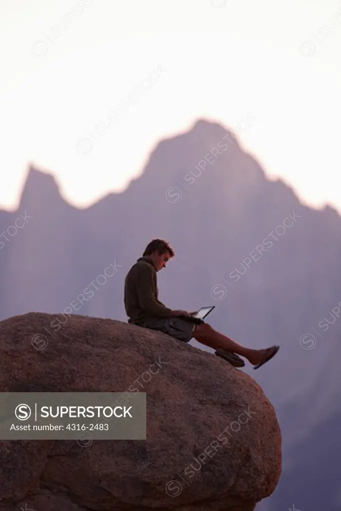 A man uses a laptop while sitting on a boulder in the Alabama Hills of California, near the town of Lone Pine. The man is sitting with Mount Whitney and surrounding peaks of the high Sierra Nevada mountains behind him.