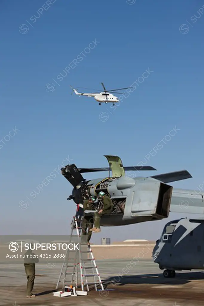An MI-17 Hip Helicopter flies past an MV-22 Osprey undergoing maintenance by U.S. Marine Corps aircraft maintainers, Camp Bastion, Helmand Province, Afghanistan.