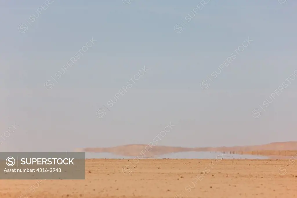 An inferior mirage dances on the horizon in the flat desert of the interior Sahara. When light rays pass from cool air to warmer air, they bend away from the direction of the temperature gradient. When air near the ground is warmer, common with hot sand or pavement, the rays bend upwards. This tricks the viewer's eye into projecting an image of the sky on the ground, which usually appears to be water or liquid in the distance.