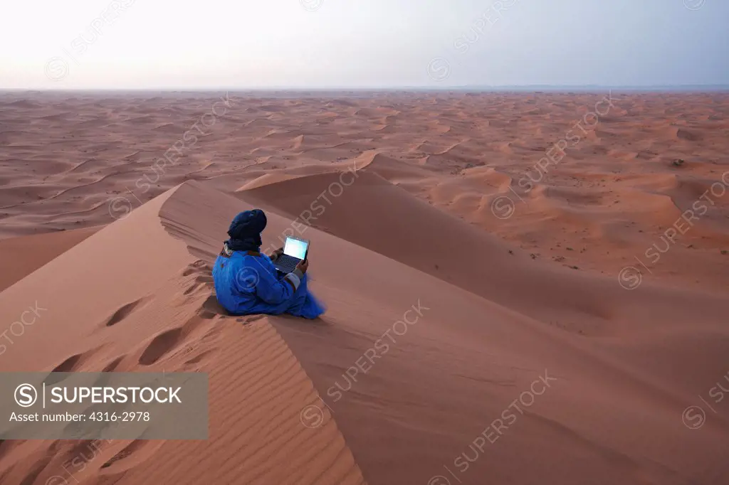 A Bedouin Nomad uses a laptop computer atop one of Morocco's highest dunes, just north of the Algerian border, interior Sahara Desert.