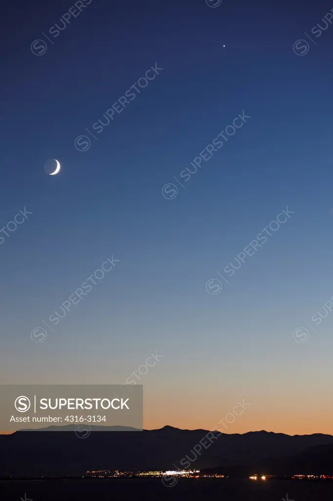 The lights of West Wendover, Nevada, as seen from the western periphery of Utah's Great Salt Lake, and a crescent moon in twilight.