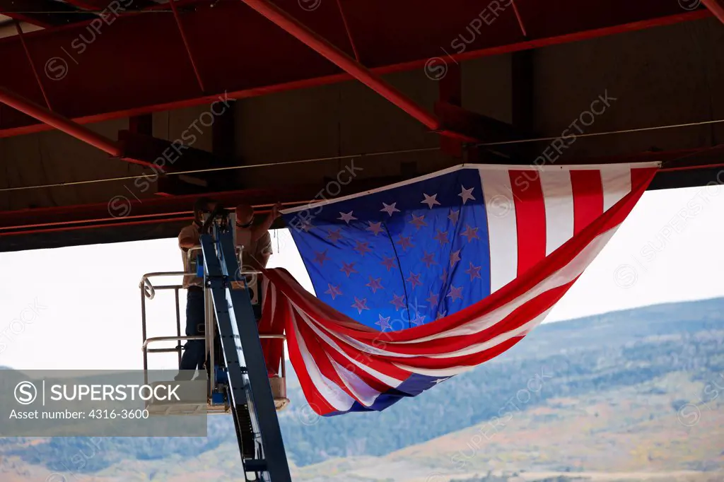 Two men on a lift folding a large American flag in an aircraft hangar.