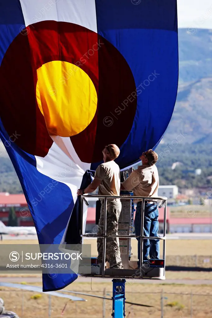 Two men on a lift folding a large State of Colorado flag.