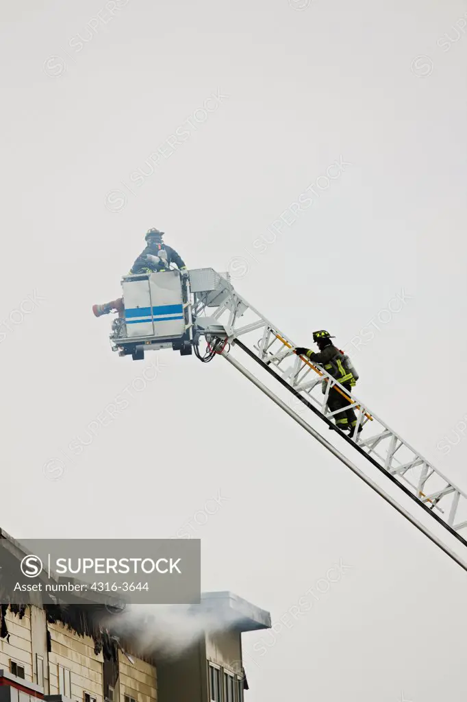 Two firefighters, one in a basket and one climbing a ladder, on a tower ladder, during a fire in a multi-story building.