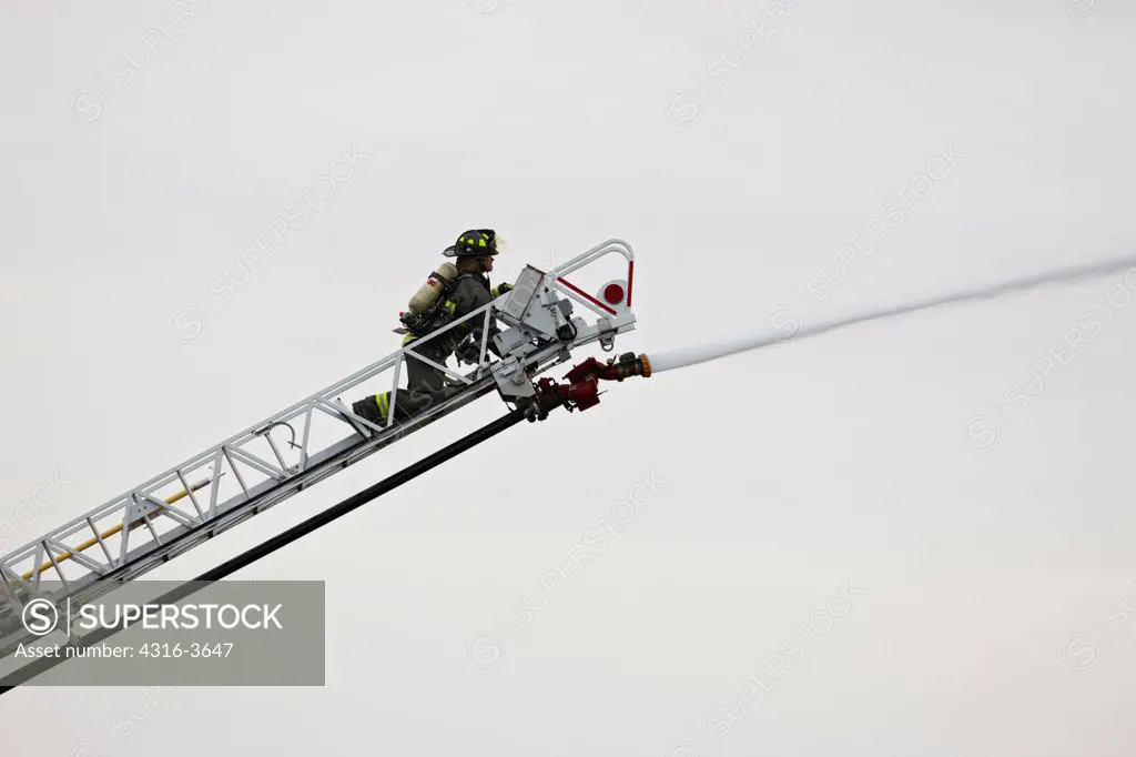 Firefighter spraying water on building fire from end of a ladder, using a water cannon.