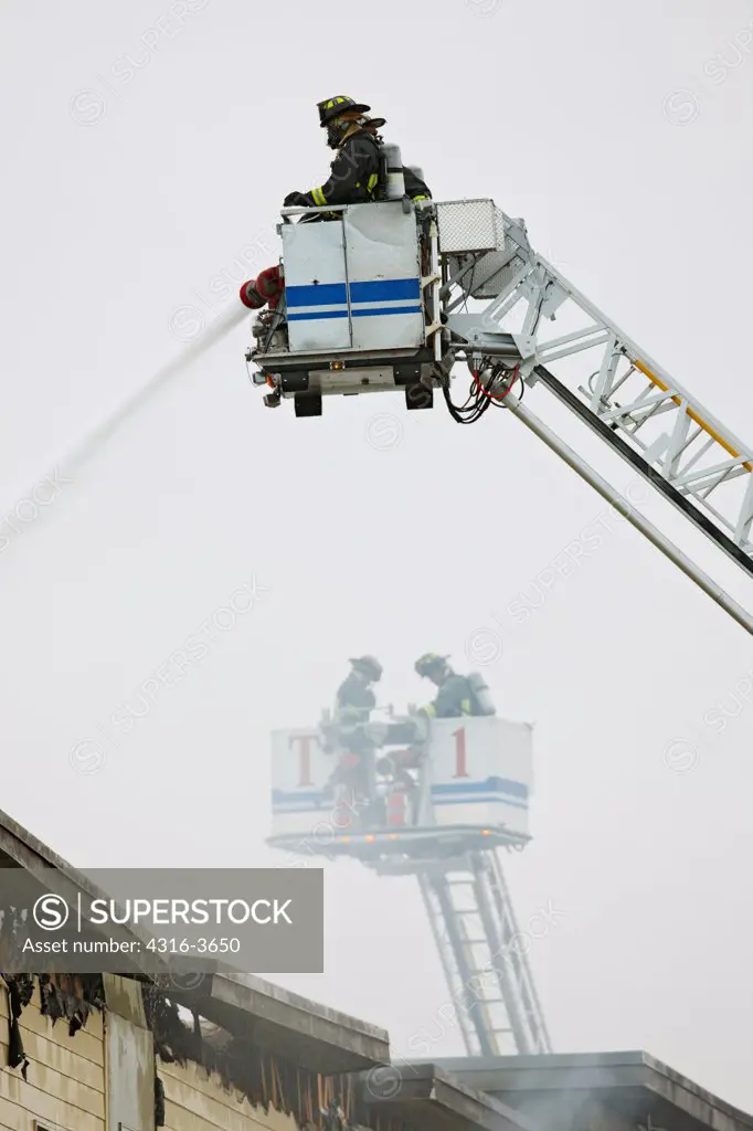 Firefighters above the fire and water damaged roof of a building, inside a basket at the end of a tower ladder.
