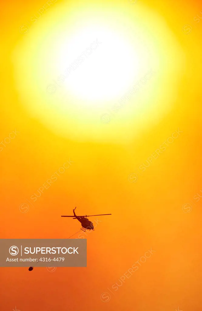 Sun silhouettes a firefighting helicopter carrying a bucket of water to a raging mountain wildfire, Colorado, USA