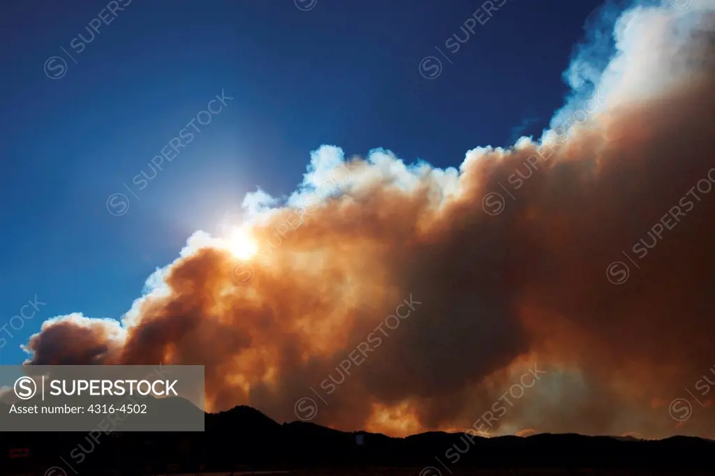 Plume of smoke rises from raging mountain wildfire, setting sun, Colorado, USA