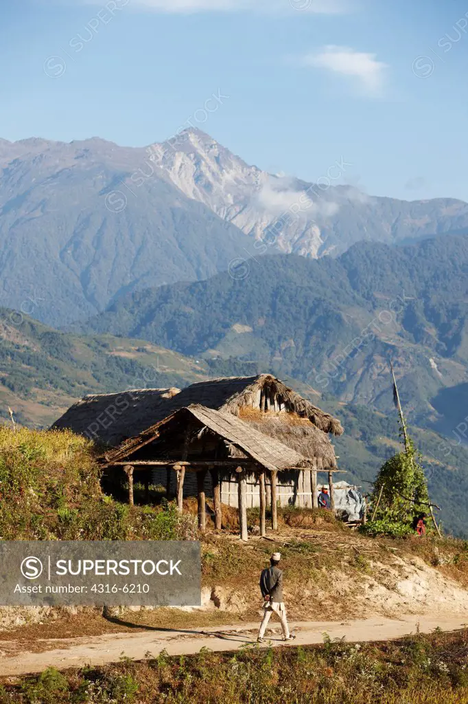 Nepal, Nepalese man walking along trail near thatch roof house and distant Himalayan peak