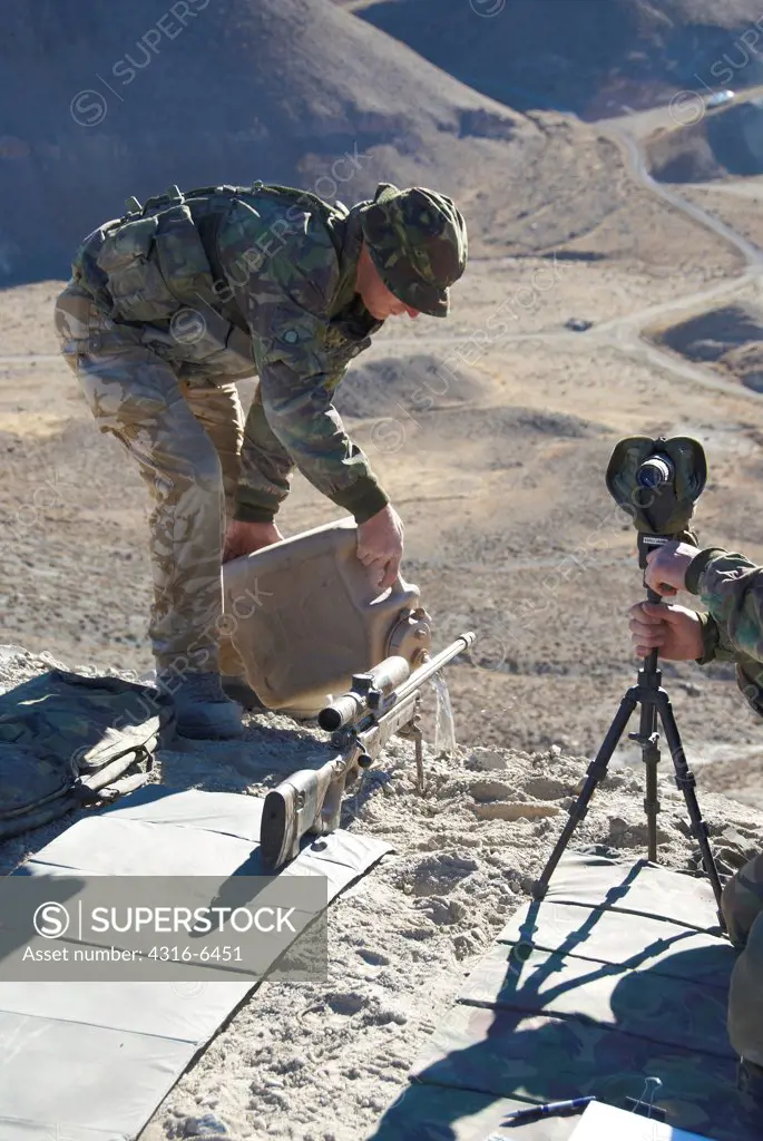 British Royal Marine pours water on dirt below the muzzle of a sniper rifle to ensure that the weapon will not lift dust into the air when it firing during mountain sniper training, Hawthorne, Nevada, USA