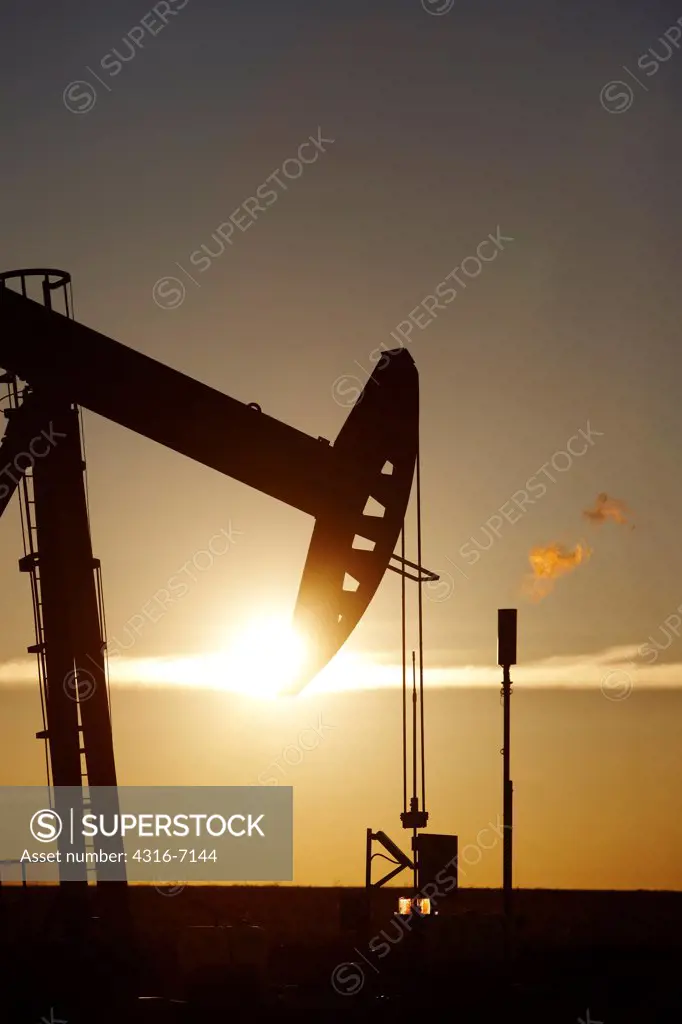 USA, Colorado, Oil well pumpjack (pump jack) and gas flare (flare stack), Oil well developed with hydraulic fracturing, also known as hydrofracking or fracking at Sunrise, diffraction of sun