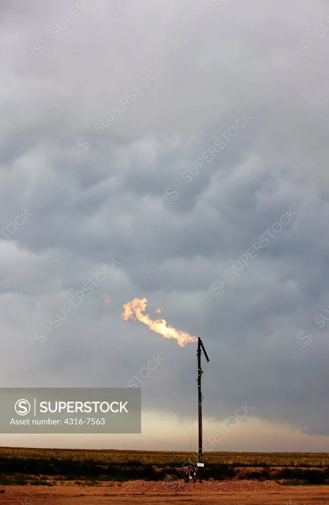 Flare stack releasing burning excess natural gas at the site of an oil well with powerful thunderstorm in distance