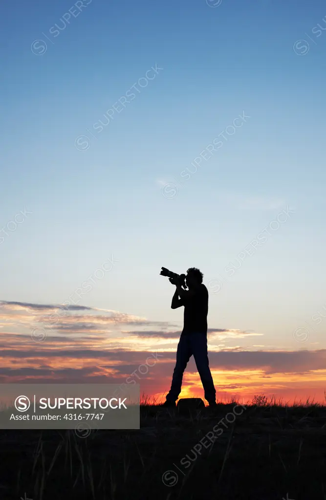 Silhouette of man aiming camera with telephoto zoom lens into sky at dawn