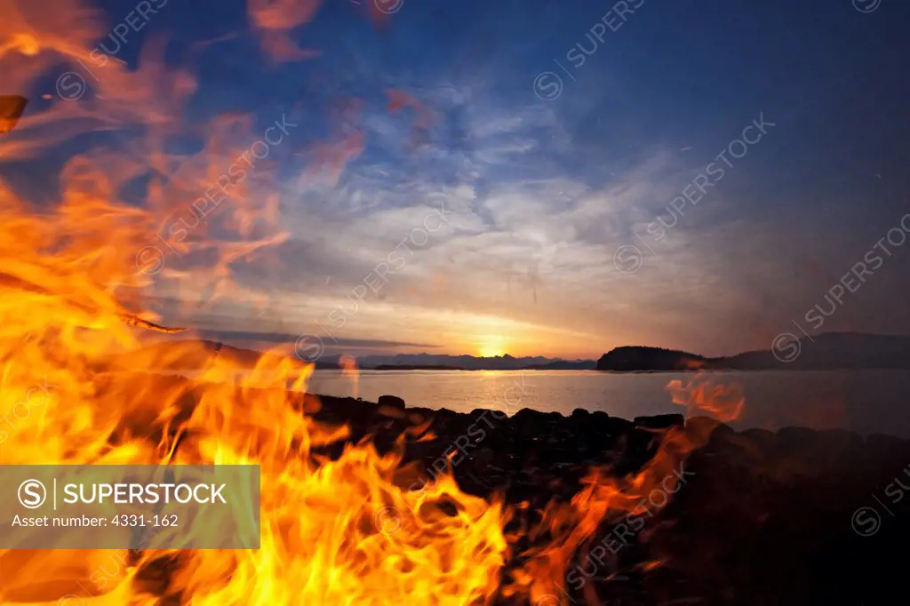 A fire on the beach on Douglas Island in Juneau, Alaska, begins to grow as a gust of wind hits. The sun sets into the Chilkat Mountain Range with Fritz Cove and Favorite Channel in between. Spuhn Island is in the foreground. The Chilkats run all the way up to Skagway.