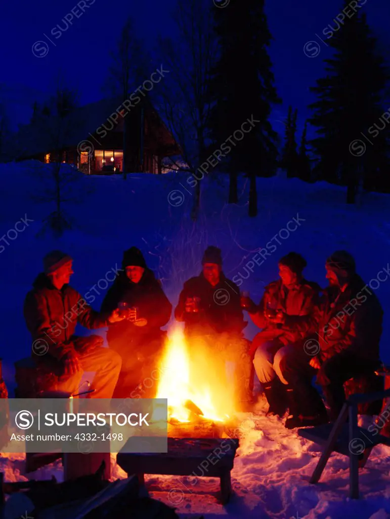 Staff and guests, Jack Jaunbral, Carl and Kirsten Dixon, Chirstina Norris, Joan Schuman and Shane Yoder, enjoying winter campfire on ice of Finger Lake, Winterlake Lodge, Alaska.