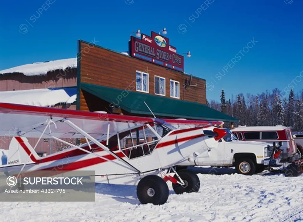 Piper PA18 Super Cub with tundra tires parked with vehicles at The Point Mackenzie General Store, Alaska.