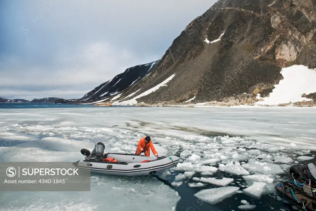 A photographer on an expedition to photograph wildlife pulls a zodiac boat over fjord ice, along a rugged coast in the Svalbard archipelago, Norway.