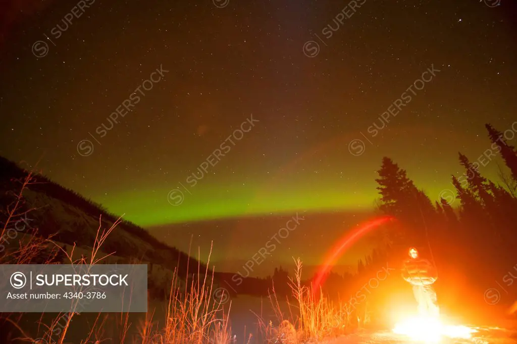 USA, Alaska, Recreational Area, Chena River State, Northern lights (Aurora borealis) glowing brightly over man enjoying hot campfire in Chena River State