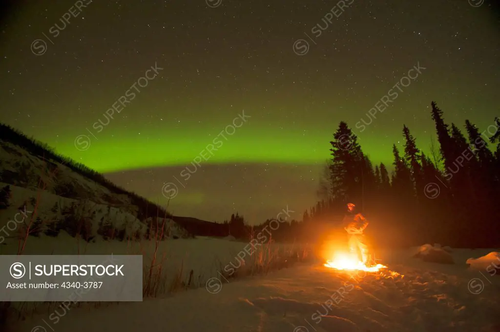 USA, Alaska, Recreational Area, Chena River State, Northern lights (Aurora borealis) glowing brightly over man enjoying hot campfire