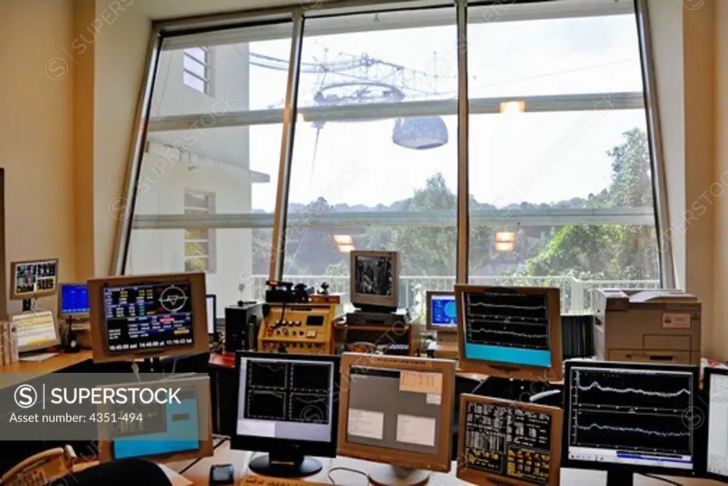 Computer monitors track observations at the Arecibo Radio Observatory.The Arecibo Observatory is a radio telescope just over 1,000 feet (305 m) across, the largest single radio telescope. The main metal collecting dish sits fixed a hemispherical karst sinkhole. The window looks out over the dish, and the spherical reflector (needed because the dish is fixed in place) can be seen hanging over the dish through the window. The observatory is the primary location of the National Astronomy and Ionosp