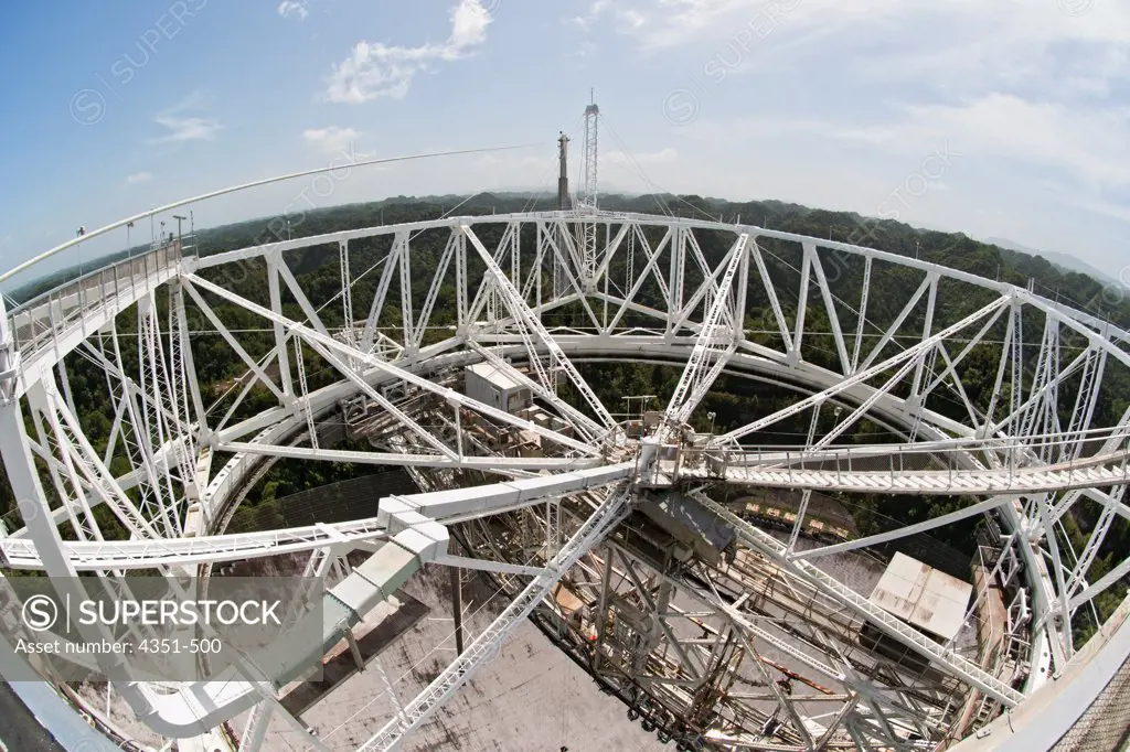 The Arecibo Observatory is a radio telescope just over 1,000 feet (305 m) across, the largest single radio telescope. The main metal collecting dish sits fixed a hemispherical karst sinkhole. Because it is fixed, a spherical reflector hangs 500 feet (150 m) over the dish on a 900 ton platform, seen here, known as 'the feed'. The observatory is the primary location of the National Astronomy and Ionosphere Center, NAIC.