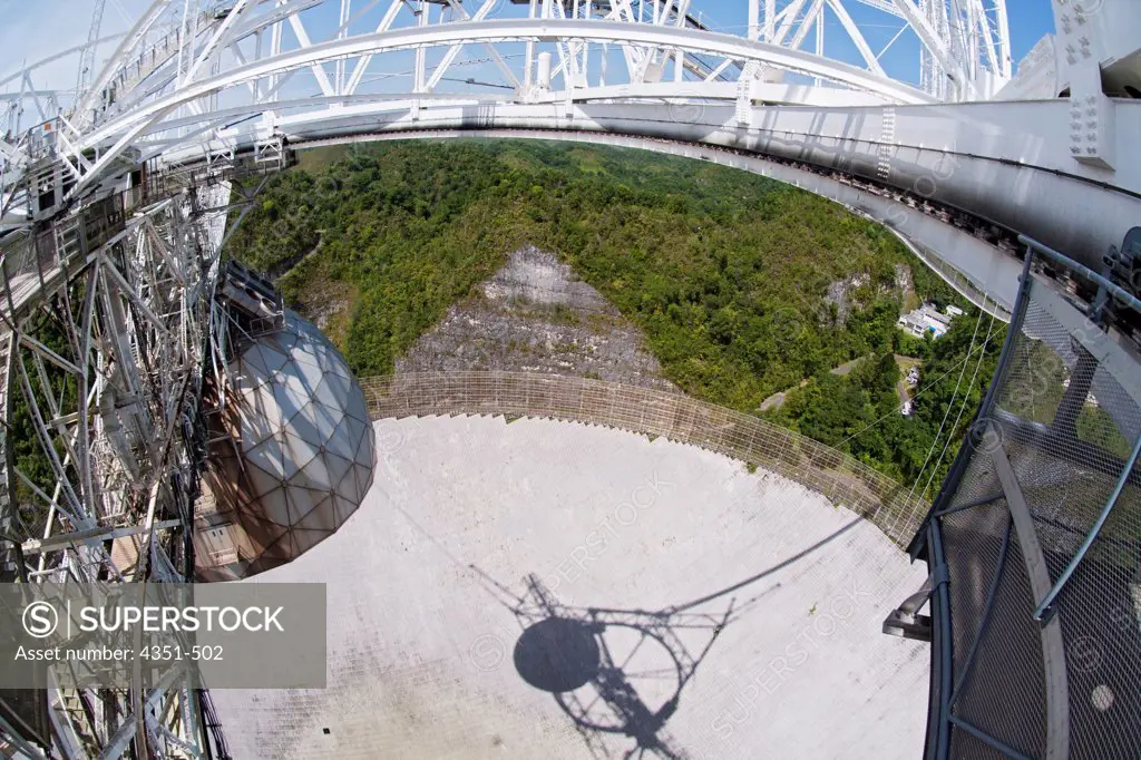 The Arecibo Observatory is a radio telescope just over 1,000 feet (305 m) across, the largest single radio telescope. The main metal collecting dish sits fixed a hemispherical karst sinkhole. Because it is fixed, a spherical reflector hangs 500 feet (150 m) over the dish on a 900 ton platform, seen here, known as 'the feed'. The observatory is the primary location of the National Astronomy and Ionosphere Center, NAIC.