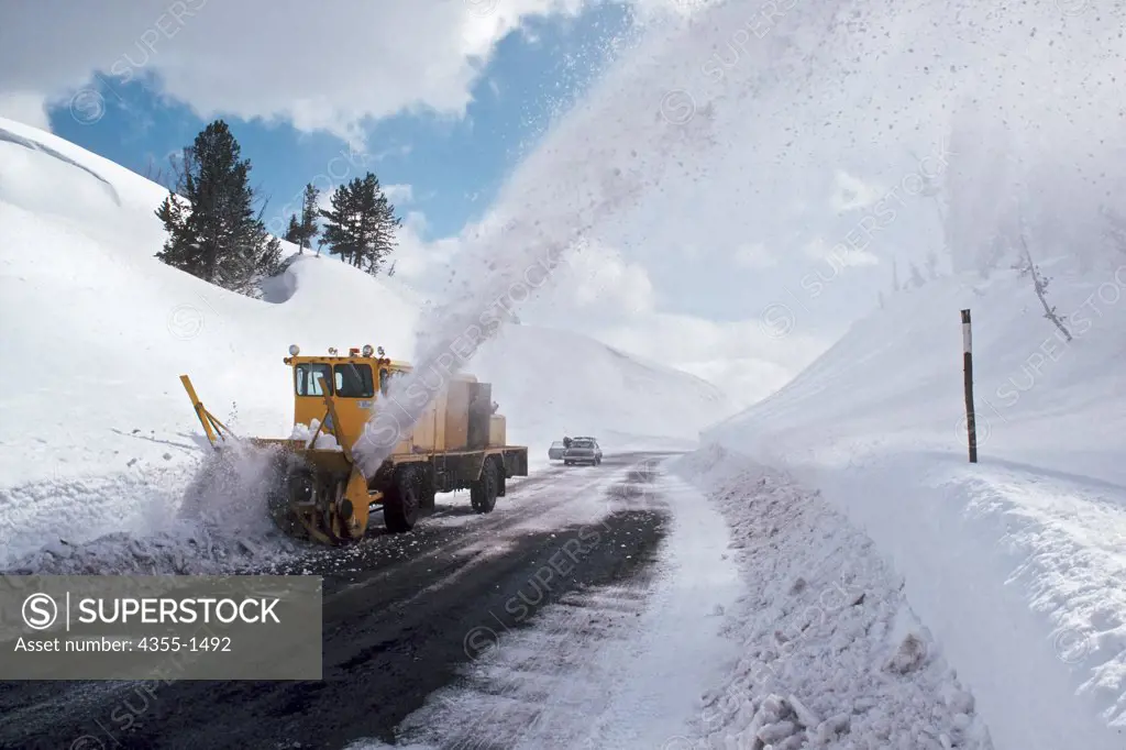 A snow removal truck uses a snowblower to remove snow from a narrow road in hilly terrain.