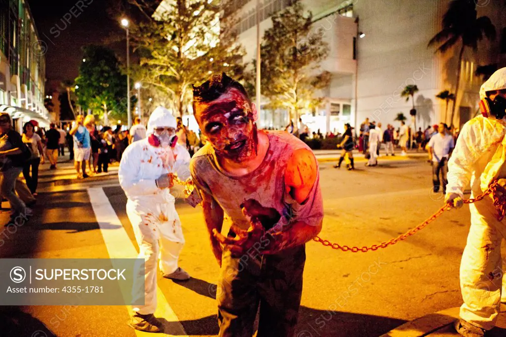 A man dressed as a zombie with haz-mat suited handlers in a Halloween event in Miami Beach.