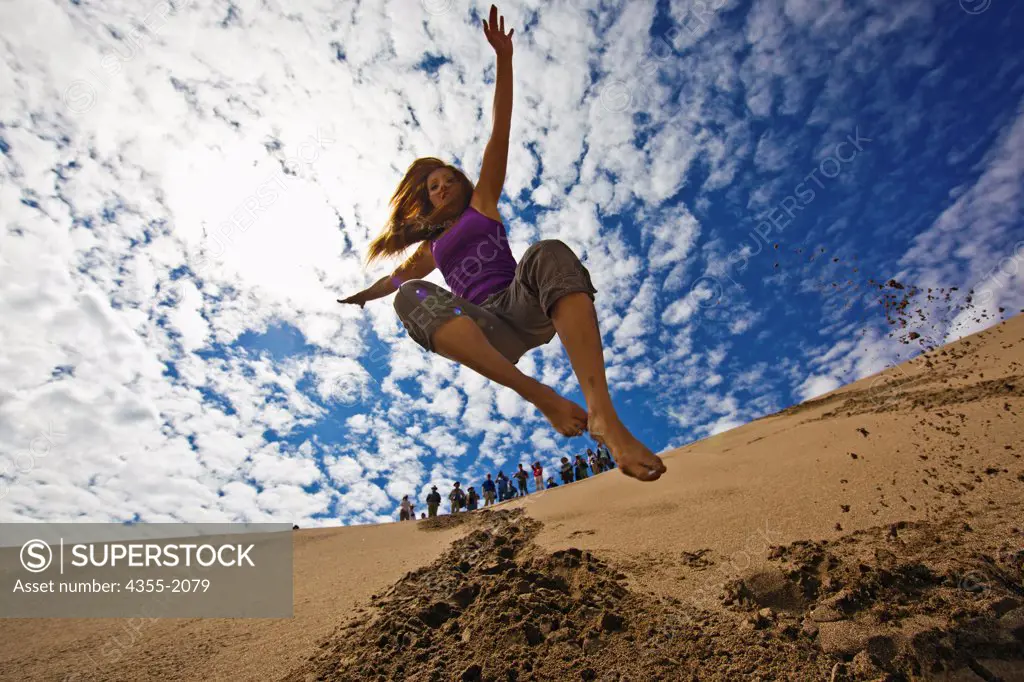 A young woman is airborne as she bounds down a sand dune while several people watch from above. Great Sand Dunes National Park and Preserve is a United States National Park located in the easternmost parts of Alamosa County and Saguache County, Colorado, United States.