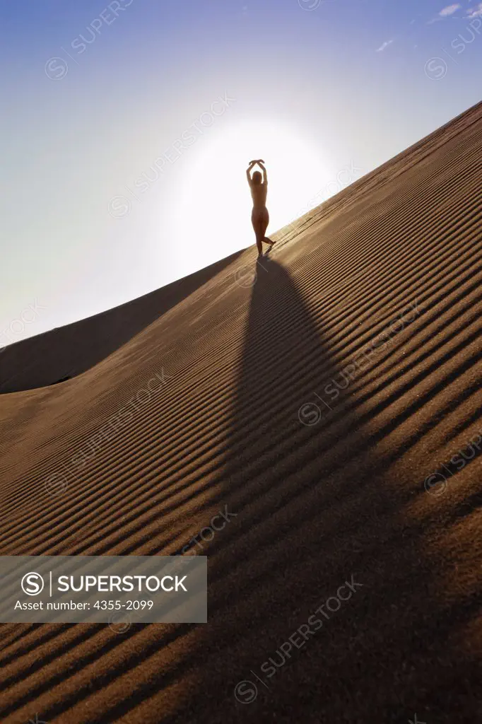 Nude young woman on sand dune at sunset.