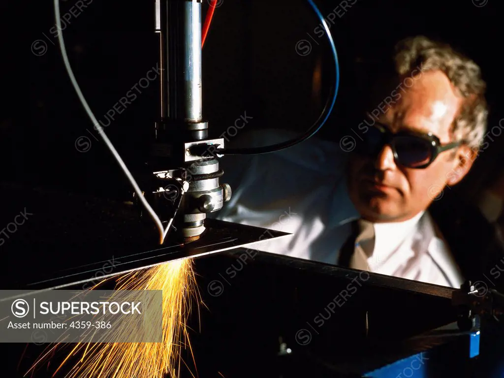A technician observes a carbon dioxide laser on a robot arm cutting a metal component in a factory.