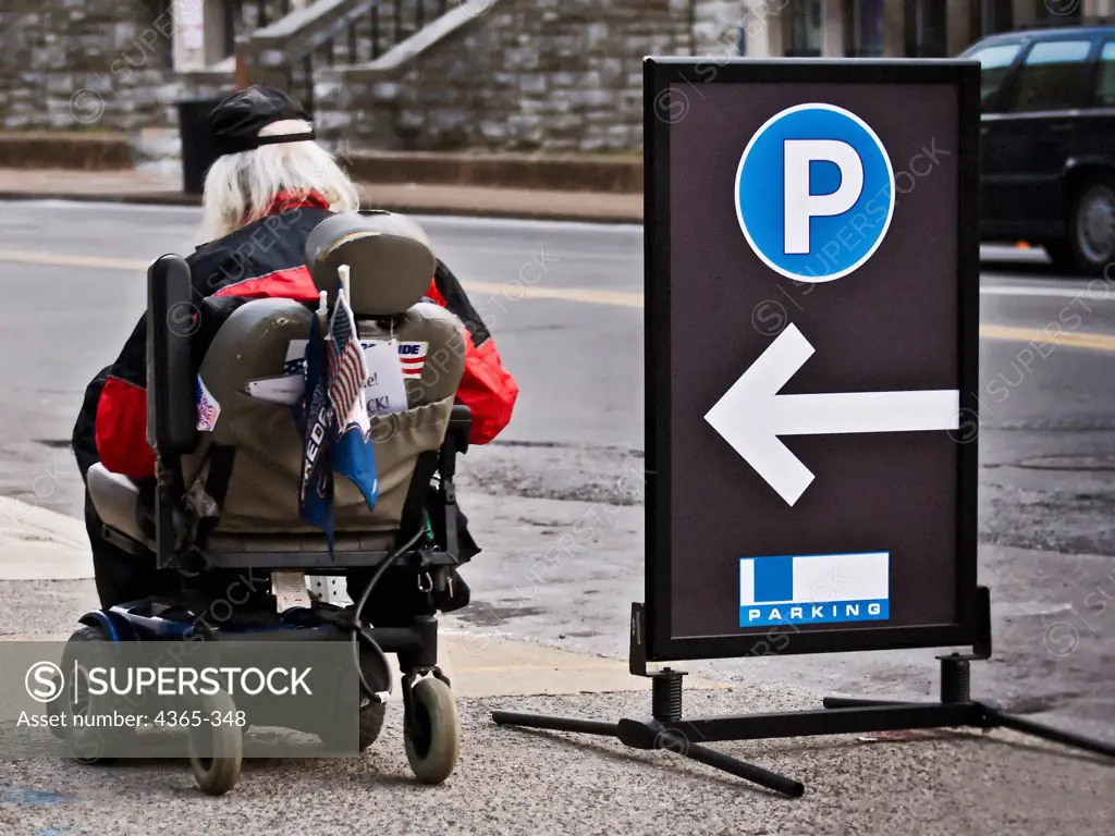 An elderly man in a motorized wheelchair rolls past a parking lot sign.