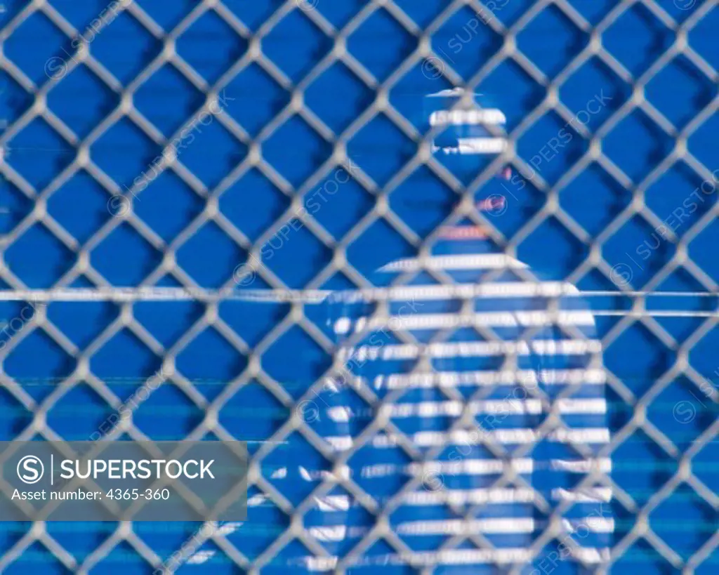 A tennis player seen through a chain-link fence and a shield.