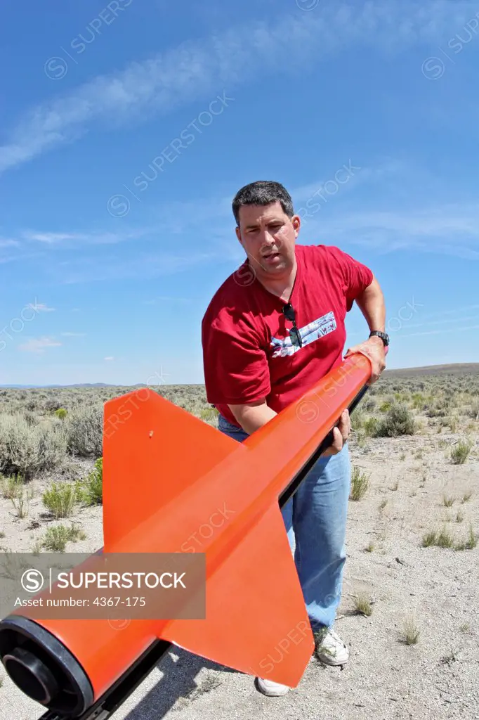 A man with the rear assembly of a red rocket (including fins and engine nozzle), at BALLS, an experimental rocketry event in the Black Rock Desert of northern Nevada.