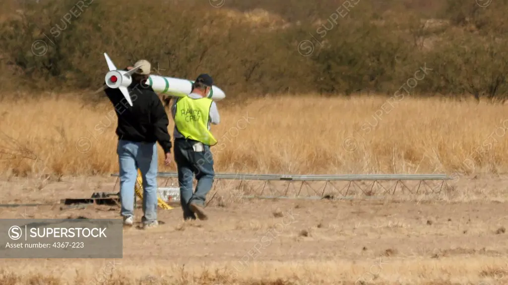 Larry Foster (in dark jacket) carries a long green and white rocket out to a launch gantry at a launch event sponsored by the Southern Arizona Rocketry Association (SARA).