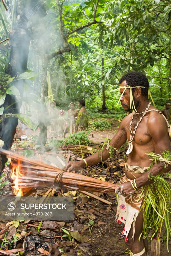 A man holds a burning bundle of palm leaves, cooking sago loaf during a village experience and cultural event, Tufi, Papua New Guinea. Sago is a starch derived from the pith of a palm tree. Participating clans were the Fighoya, Kandoro, Tewari, Gaboru, and Safu from the villages of Koje, Kofure, Iagirua, Koave, Marabade, Fodoma, Bekoyana, Koyatona.