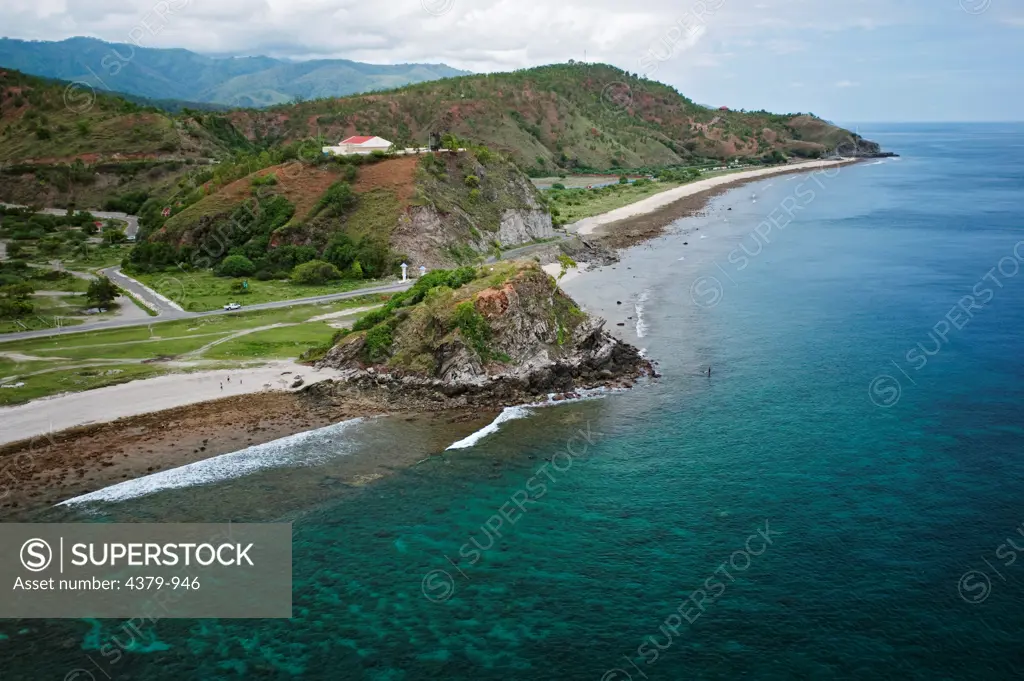 An aerial view of the coast west of Dili, East Timor. The rock and posts on the road mark the western end of Dili, East Timor's capital. The red-roofed building on the hill  has a large statue of Pope John Paul II.
