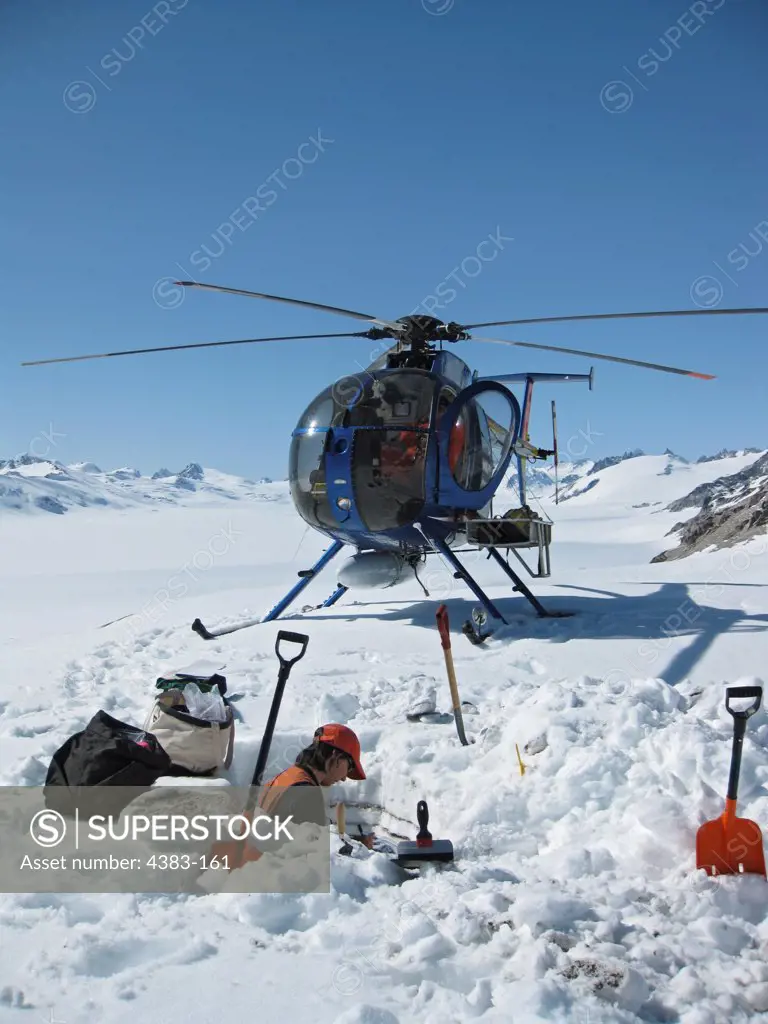 Geologist Kristi Wallace sampling tephra deposits preserved in the snow north of Redoubt volcano after digging through alternating layers of snow and tephra in front of her. The helicopter which brought her to the site is parked in front of her. Image by Janet Schaefer for the Alaska Volcano Observatory/U.S. Geological Survey.